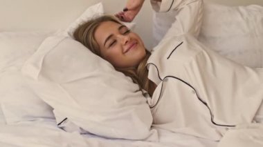 Pijama giyen genç, mutlu bir sarışın kız sabah yatak odasında uyanıyor.