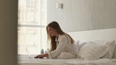 Pijama giymiş gülümseyen kadına bakan kamera dizüstü bilgisayarında daktilo yazarken ışık odasındaki yatağın üzerine oturmuş notlar alıyor.