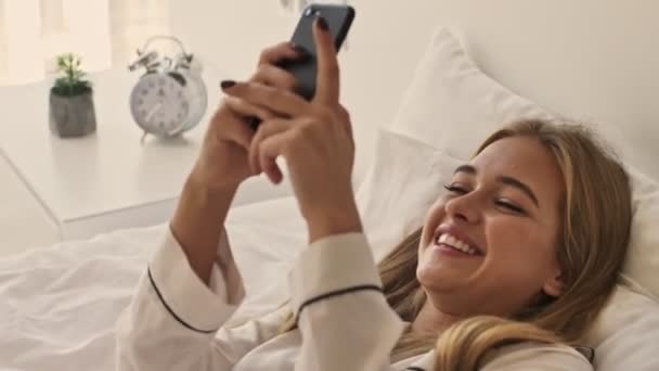 Pijama Giyen Gülümseyen Sarışın Bir Kız Yatakta Akıllı Telefon Kullanıyor — Stok video