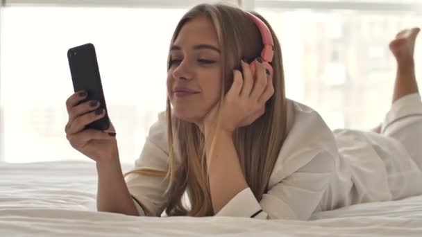 파자마를 귀여운 금발의 침대에서 스마트폰을 사용하면서 헤드폰으로 음악을 — 비디오