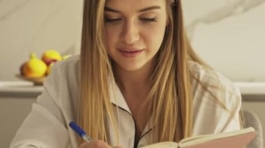 Pijamalı güzel, gülümseyen genç bir kadın sabah mutfakta dizüstü bilgisayarını kullanırken defterine bir şeyler yazıyor.