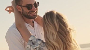 Mutlu ve mutlu çiftin günlük kıyafetleri içinde güneşli kumsalda birbirlerine bakarken kucaklaşıyorlar.