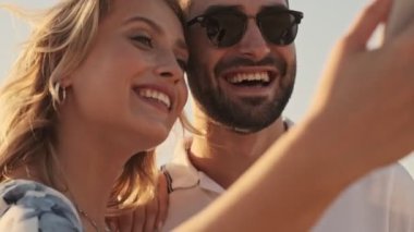 Gündelik giysiler içinde gülümseyen genç bir çiftin yakın çekim görüntüsü güneşli sahilde selfie çekiyor.