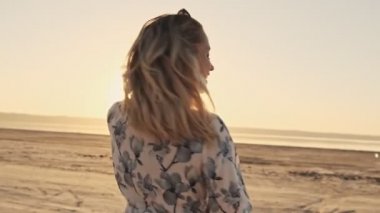 Güzel sarışın bir kız kameraya doğru dönüyor ve gölün yanındaki güneşli sahilde göz kırpıyor.