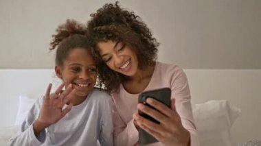Neşeli Afrikalı kadın ve küçük güzel kızı akıllı telefondan video konuşmaları yapıyorlar ve evde yatakta otururken kameraya el sallıyorlar.