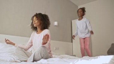 Memnuniyet duyan Afrikalı bir kadın, küçük kızı yatakta arkasında zıplarken gözleri kapalı meditasyon yapmaya çalışıyor.