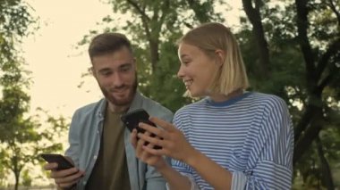Genç gülümseyen kız, parkta bir bankta otururken akıllı telefonuyla erkek arkadaşına bir şey gösteriyor.