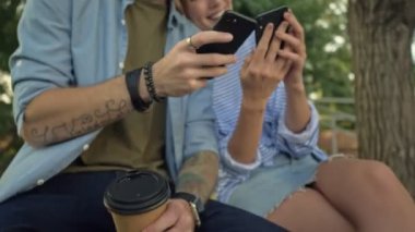 Genç ve gülümseyen bir çiftin yakın çekim görüntüsü, park manzarasında elektrikli scooterlarla bankta otururken akıllı telefonlar kullanıyor.