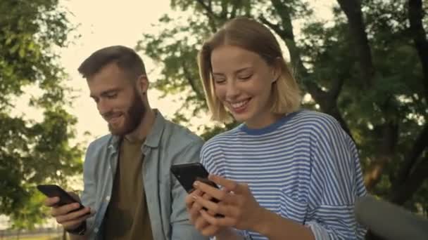 Unga Leende Par Använder Smartphones Medan Sitter Bänken Park Landskap — Stockvideo