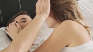 Çekici, birbirine aşık genç bir çift, beyaz bir yatakta uzanıyorlar ve birbirlerine sarılıp konuşuyorlar.