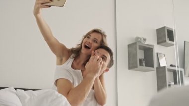 Mutlu gülümseyen genç bir çift sabah beyaz bir yatak odasında selfie çekiyor.