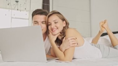 Memnun genç bir çift sabah beyaz bir yatak odasında dizüstü bilgisayar kullanıyor.