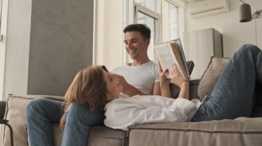 Gülümseyen genç bir çift erkek arkadaş ve kız arkadaş akıllı telefon kullanarak ve oturma odasında kitap okuyarak vakit geçiriyorlar.