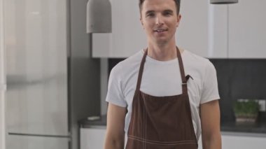 Kahverengi önlük giyen gülümseyen genç adam, başparmağını mutfaktaki kameraya doğru gösteriyor.