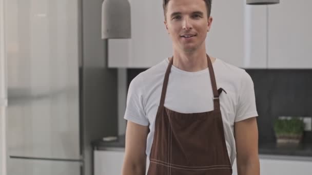 茶色のエプロンを身に着けている笑顔若い男は キッチンでカメラに親指を表示しています — ストック動画