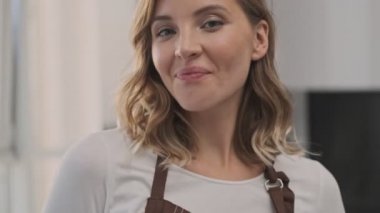 Sevimli genç sarışın bir kadın kameraya gülümsüyor. Kapalı mekanda yüzüne dokunurken.