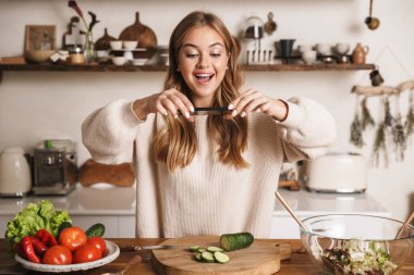 Heyecanlı, sıradan kıyafetler giyen, cep telefonuyla fotoğraf çeken ve rahat bir mutfakta gülümseyen güzel bir kadın resmi.
