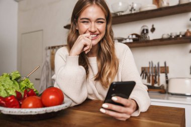 Kafkasyalı neşeli bir kadının rahat bir mutfakta yemek pişirirken cep telefonu kullanması.