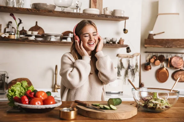 부엌에서 식사를 준비하면서 헤드폰을 사용하여 평상복을 미소짓는 여성의 — 스톡 사진