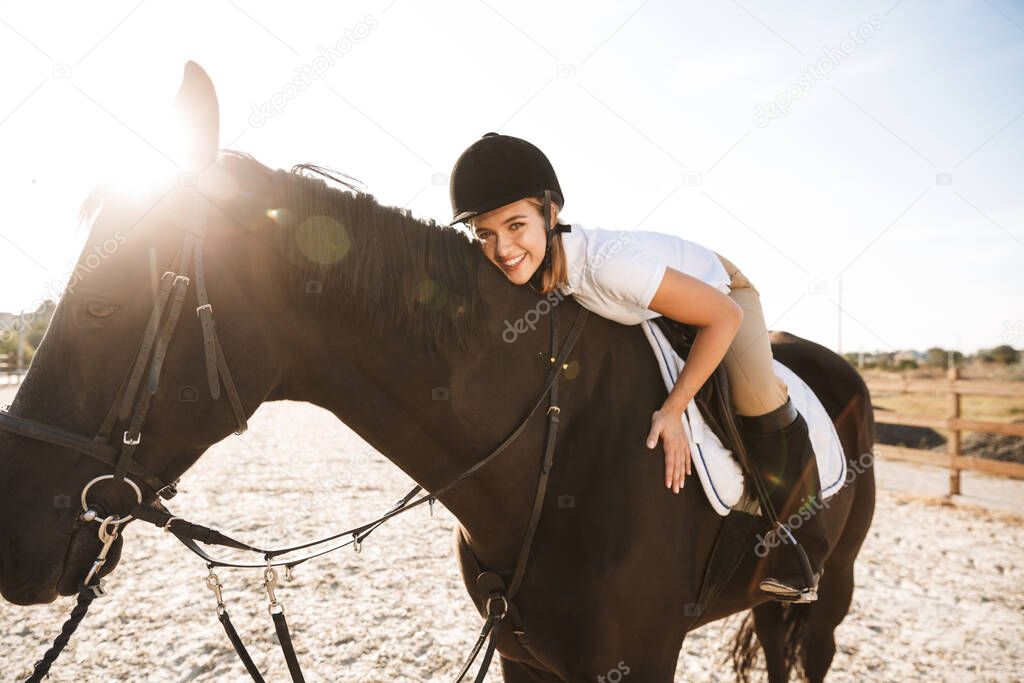 Image of cheery optimistic young blonde beautiful woman wearing hat with horse in countryside outdoors