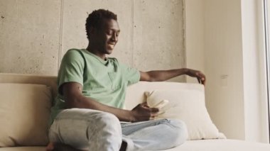 Tatmin olmuş Afro-Amerikan bir adam evde kanepede otururken akıllı telefondan bir şey izliyor.