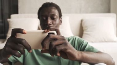 Sıradan giyinmiş çekici bir Afro-Amerikan erkeği akıllı telefonunu oturma odasında yatay olarak tutarken kullanıyor.