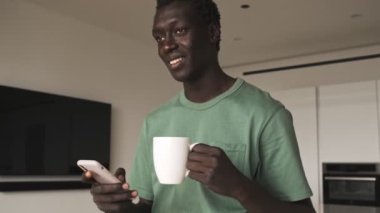 Gülümseyen Afrikalı Amerikalı bir adam evinde elinde çay tutarken akıllı telefonunu kullanıyor.