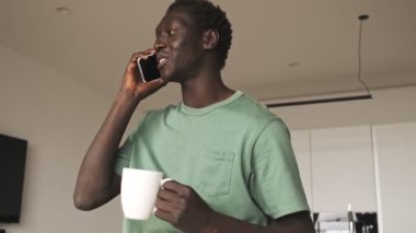 Olumlu bir Afro-Amerikan erkeği evinde elinde çay tutarken akıllı telefonuyla konuşuyor.