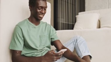 Mutlu, gülen Afro-Amerikan bir adam, günlük giysiler içinde oturma odasında akıllı telefonundan bir şey izliyor.