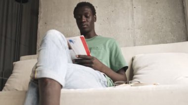 Rahat giyinmiş Afro-Amerikan bir adam oturma odasındaki kanepede oturmuş dergi okuyor.