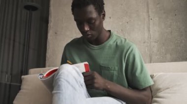 Ciddi bir Afro-Amerikalı günlük giysiler içinde oturma odasındaki kanepede oturan bir kitaba notlar yazıyor.