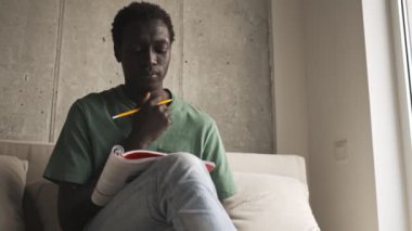 Odaklanmış Afro-Amerikan bir adam günlük giysiler içinde oturma odasındaki kanepede oturan bir kitapta görevler yapıyor.