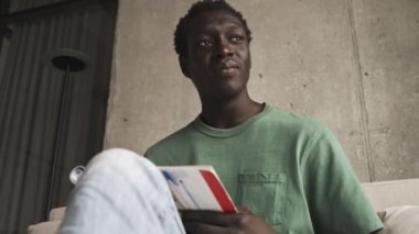 Günlük giysiler içinde yorgun bir Afrikalı Amerikalı oturma odasındaki bir kitapta görevlerini yaparken yan tarafa bakıyor.