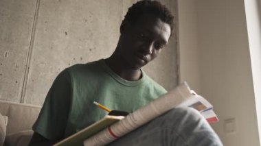 Odaklanmış, ciddi bir Afro-Amerikalı, günlük giysiler içinde oturma odasındaki bir kitapta alıştırma yapıyor.