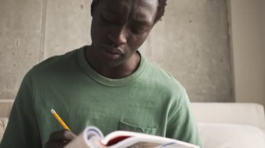 Sakin, ciddi, Afro-Amerikan bir adam gündelik giysiler içinde oturma odasındaki bir kitapta alıştırma yapıyor.