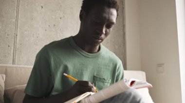 Sakin odaklı Afro-Amerikan bir adam gündelik giysiler içinde ödevlerini oturma odasında yaparken bir kitaba not alıyor.