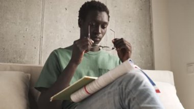 Sakin ve yakışıklı Afro-Amerikan bir adam, günlük giysiler içinde oturma odasında kitap okurken gözlüğünü takıyor.
