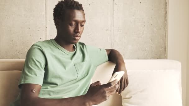 一位穿着休闲装的自信的非洲裔美国人坐在家里的沙发上 一边用他的智能手机 — 图库视频影像