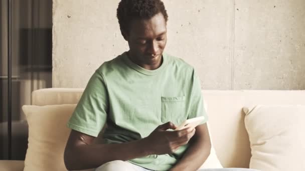 一位穿着休闲装的冷静的非洲裔美国人坐在家里的沙发上 一边用他的智能手机 — 图库视频影像