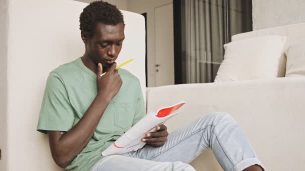 一位穿着休闲装的沉思的非洲裔美国人一边在客厅里拿着一本书和铅笔一边学习 — 图库视频影像