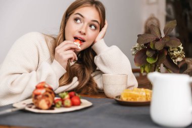 Kafkasyalı hoş bir kadının rahat bir odada kahvaltı ederken günlük giysiler giyip çilek yemesi.