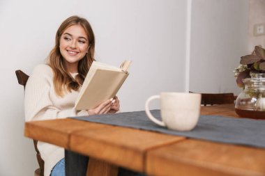 Sıradan kıyafetler giyen, kitap okuyan ve rahat bir odada çay içen beyaz bir kadın resmi.