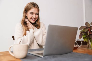 Heyecanlı genç bir kadının rahat bir odada çay içmesi ve dizüstü bilgisayar kullanması.