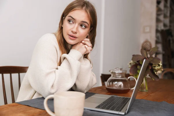 若い白人女性がノートパソコンを使ってカジュアルな服を着て居心地の良い部屋でお茶を飲む姿のイメージ — ストック写真