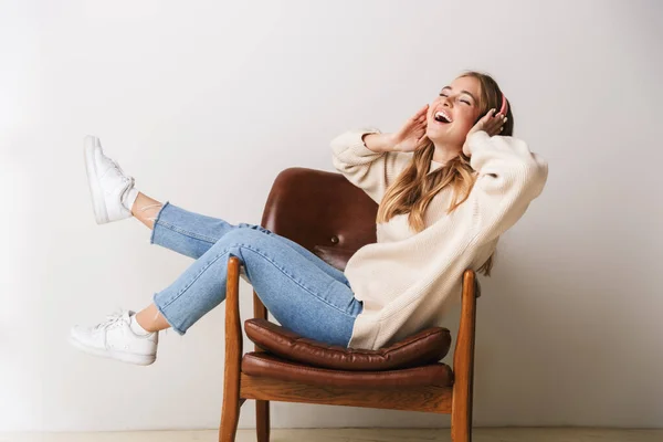 兴奋的年轻女子坐在白色背景的扶手椅上 面带微笑 使用耳机的照片 — 图库照片