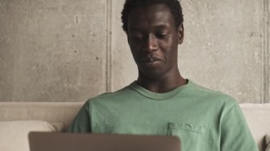 Mutlu, Afro-Amerikan bir adam oturma odasında gümüş dizüstü bilgisayarını kullanırken gülümsüyor.