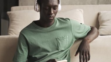 Kablosuz kulaklıklı sakin, Afro-Amerikalı bir adam oturma odasında oturmuş akıllı telefonunu kullanarak müzik dinliyor.