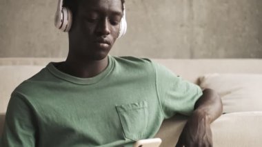 Kablosuz kulaklıklı Afro-Amerikan bir adam oturma odasında akıllı telefonunu kullanırken müzik dinliyor.