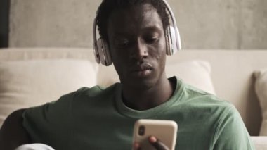 Kablosuz kulaklıklı mutlu bir Afro-Amerikalı adam müzik dinliyor ve evdeki oturma odasında gülümsüyor.