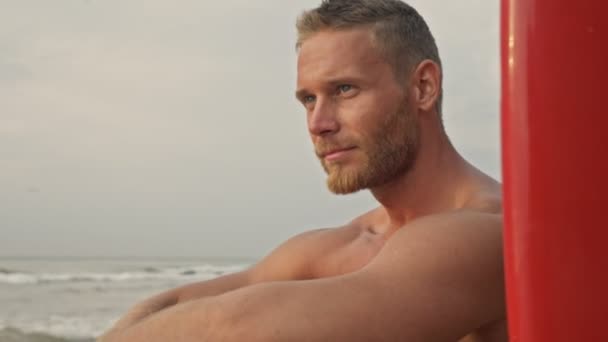 笑顔ひげを生やしたハンサムな男離れて見ている間に座ってサーフボードで海の近くのビーチ — ストック動画